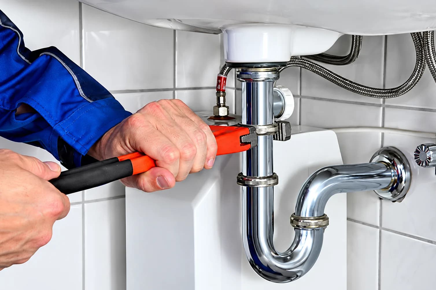 A plumber tightening a chrome trap beneath a basin with a pipe wrench